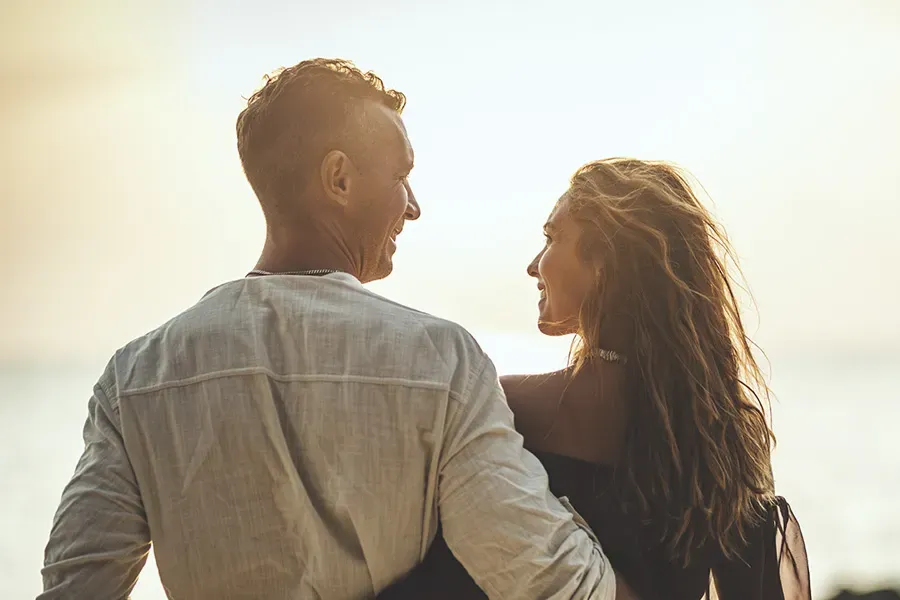 A couple gazes at each other on the beach at sunrise, viewed from behind. Schedule Anti-Aging treatment from Morgan Burns, APRN in New Smyrna Beach.