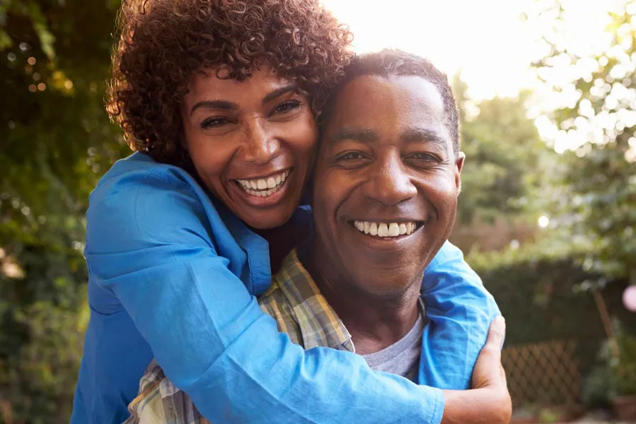 A middle-aged African American couple outside, the woman embracing the man from behind pressing her cheek to his, both are smiling due to successful Autoimmune Disorder treatment from Morgan Burns, APRN in New Smyrna Beach.