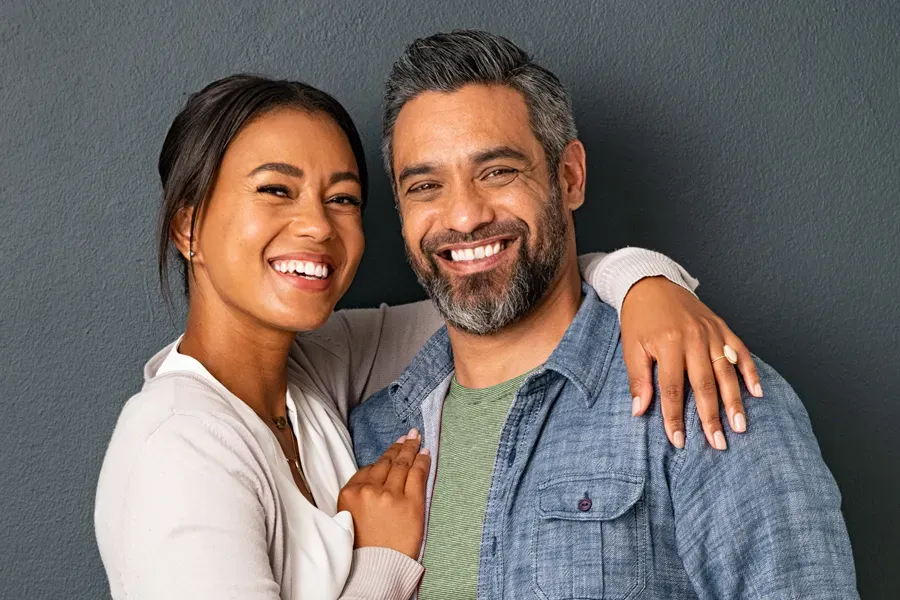 A middle-aged couple with their arms around each other standing against a gray background and smiling, happy with the bioidentical hormone therapy they received from Morgan Burns, APRN in New Smyrna Beach.