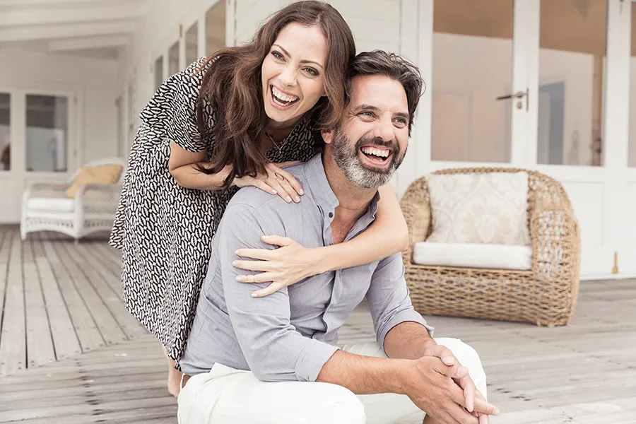 A middle-aged couple on a wooden porch with wicker furniture; the woman is playfully hugging the man from behind. They are benefiting from the concierge care services from Morgan Burns, APRN in New Smyrna Beach.