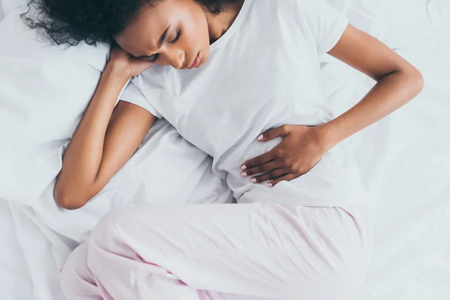 A dark-skinned woman in white clothes lays on her bed clutching her middle before getting treatment for Crohn's Disease from Morgan Burns, APRN in New Smyrna Beach.