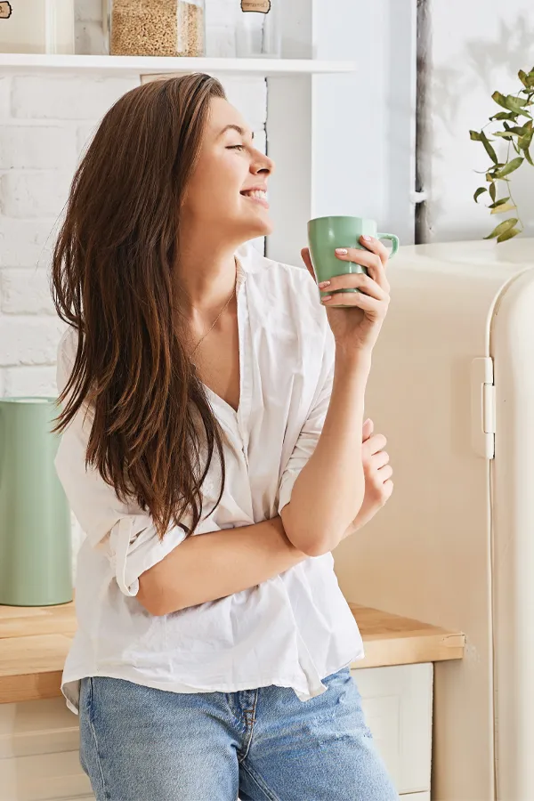 A woman with a white blouse an jeans drinking tea in her kitchen, smiling after getting treatment for Crohn's Disease from Morgan Burns, APRN in New Smyrna Beach.