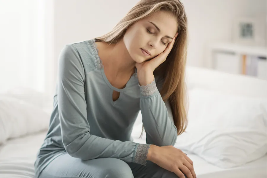 A woman sits on the edge of her bed with her head in her hands. Schedule treatment for depression from Morgan Burns, APRN in New Smyrna Beach.