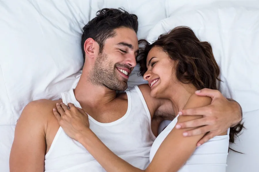 A laughing man and woman in undershirts lie in each other's arms on a white bed, representing sexual health peptide therapy by Morgan Burns, APRN in New Smyrna Beach.