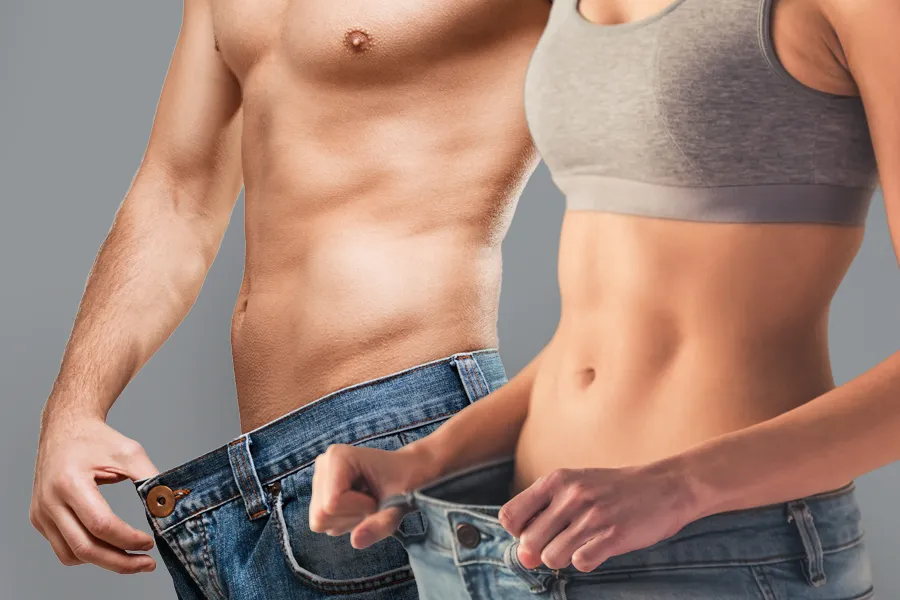A man and a woman with toned bodies hold their jeans away from their waists to show weight loss. Get weight loss peptide treatment from Morgan Burns, APRN in New Smyrna Beach.