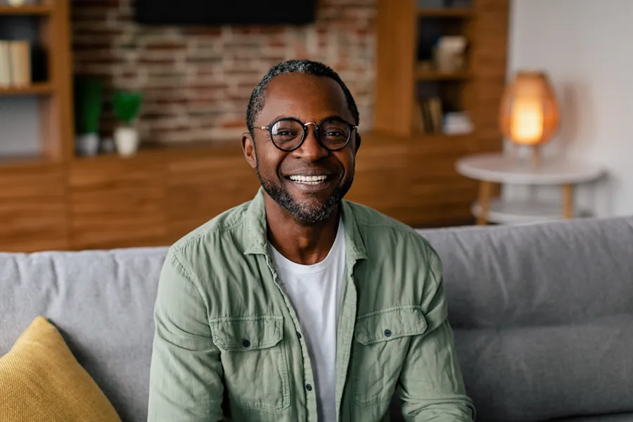 A middle aged black man in a green button up shirt sits on his couch smiling, happy with his treatment for andropause and low testosterone from Brandi Terra, NP at Oak & Stone Wellness in San Diego.