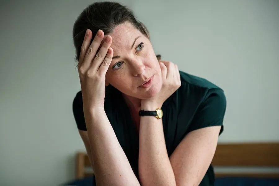 A woman in a dark blue dress sits on her bed holding her head in her hands with an exhausted expression. Get help for chronic fatigue syndrome from Brandi Terra, NP at Oak & Stone Wellness in [AERA].