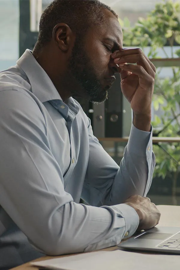A black man rubbing the top of his nose at his desk, in need of chronic fatigue treatment from Brandi Terra, NP at Oak & Stone Wellness in San Diego.