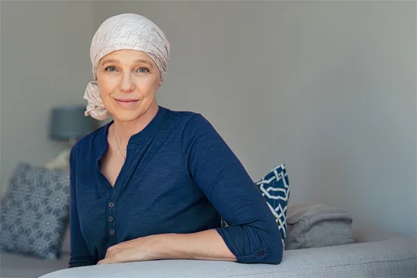 A woman wearing a blue shirt with a white scarf tied around her head sitting on a couch and smiling at the camera, representing sensitive functional oncology treatment from Brandi Terra, NP at Oak & Stone Wellness in San Diego.