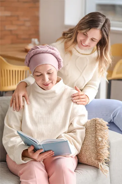 A woman with a scarf tied around her head sits on a couch reading while another woman sits behind her with her hands on her shoulders warmly, representing sensitive functional oncology treatment from Brandi Terra, NP at Oak & Stone Wellness in San Diego.