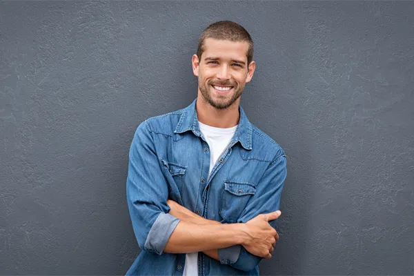 A man in a denim shirt stands smiling against a gray-blue wall, pleased with his testosterone hormone therapy from Brandi Terra, NP at Oak & Stone Wellness in San Diego.