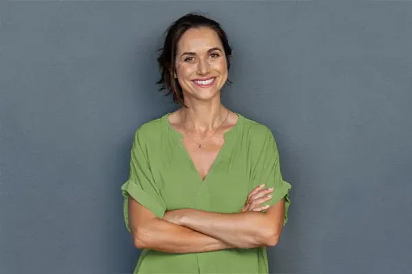 A woman in a green shirt stands smiling against a gray-blue wall, pleased with her hormone therapy from Brandi Terra, NP at Oak & Stone Wellness in San Diego.