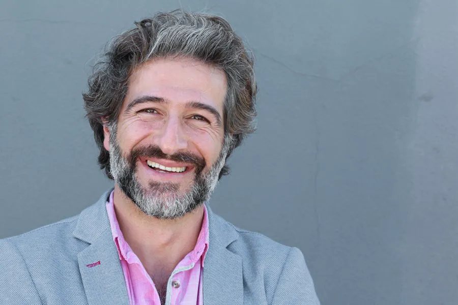 A smiling man with salt and pepper hair and beard wearing a pink shirt and gray blazer stands agains a gray wall. Get hypogonadism treatment from Brandi Terra, NP at Oak & Stone Wellness in San Diego.
