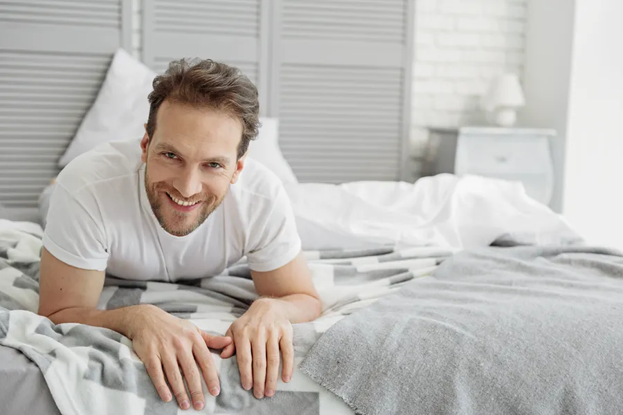 A dirty-blonde haired man lays on his stomach in bed smiling. Schedule treatment for low libido in men from Brandi Terra, NP at Oak & Stone Wellness in San Diego.
