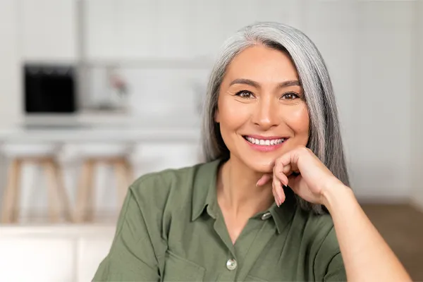 An asian woman with long gray hair and a green shirt sitting and smiling on her couch, happy with her menopause care from [NAME} in San Diego.