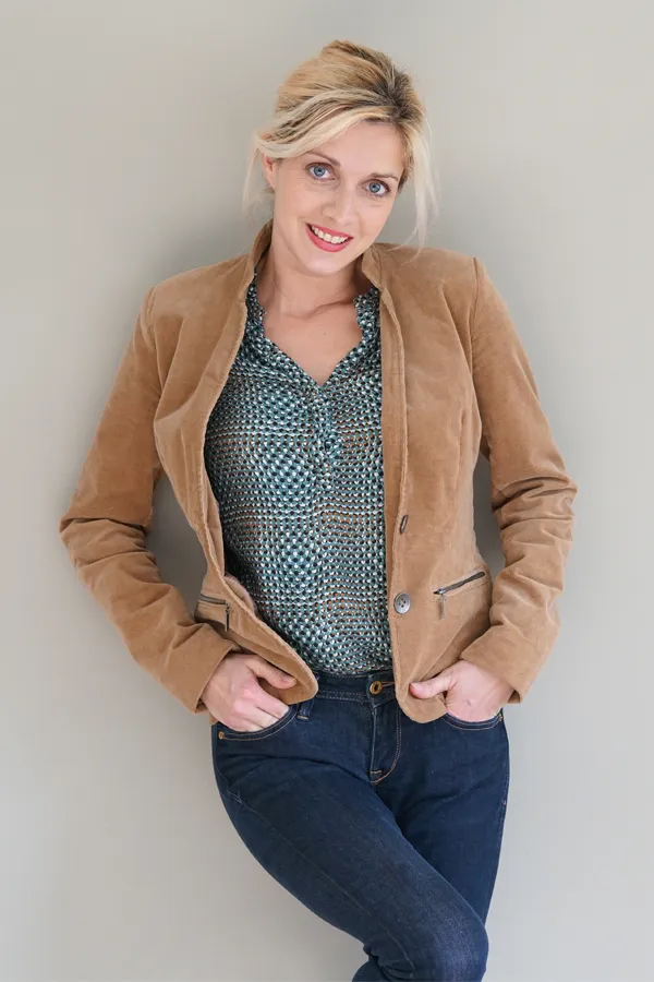 A smiling blonde woman in a tan leather jacket leans against a gray wall. Schedule metabolic syndrome from Brandi Terra, NP at Oak & Stone Wellness in San Diego.
