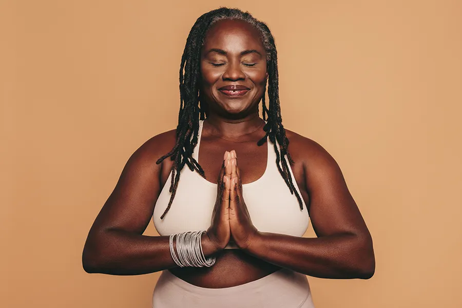 An older African American woman confidently posing against a warm background smiling with hands in a prayer pose. Brandi Terra, NP at Oak & Stone Wellness treats obesity using functional medicine in San Diego.