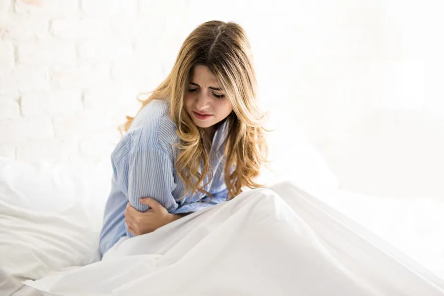 A woman with long blonde hair sits in her bed holding her stomach in pain, considering getting care for PMS from Brandi Terra, NP at Oak & Stone Wellness in San Diego.