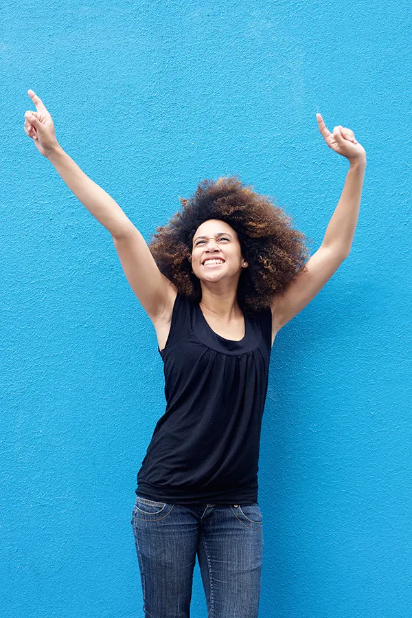 A woman in a dark blue tank top standing in front of a bright blue wall, raising her arms in celebration of relief from PMS from Brandi Terra, NP at Oak & Stone Wellness in San Diego.