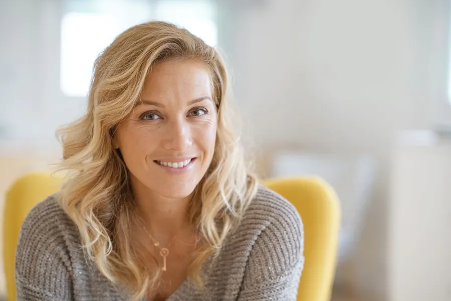 A blonde woman wearing a a gray sweater sits inside in a yellow chair, smiling and happy with her perimenopause treatment from Brandi Terra, NP at Oak & Stone Wellness in San Diego.