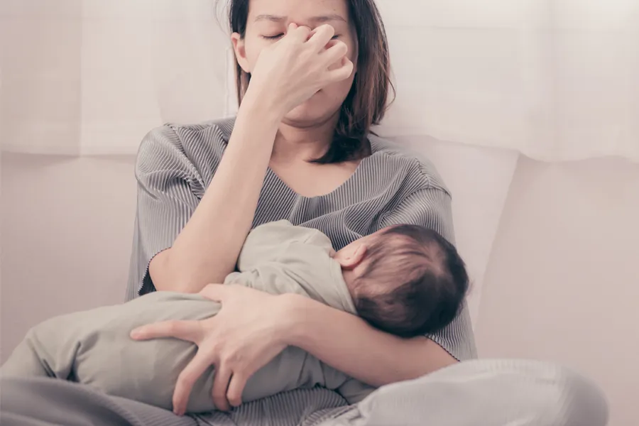 A woman nursing an infant on couch rubs her eyes with exhaustion. Get treatment for postpartum hormone imbalance from Brandi Terra, NP at Oak & Stone Wellness in San Diego.