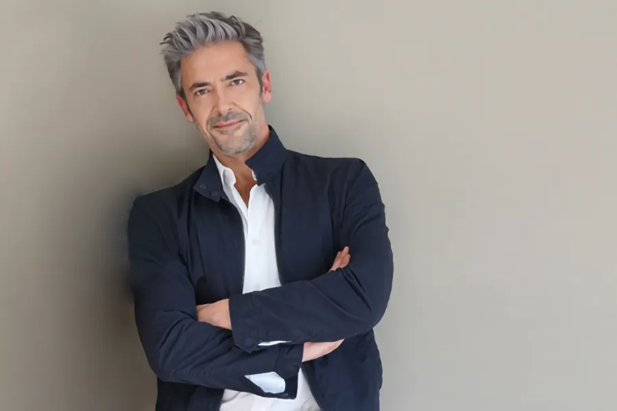 A middle-aged man in a white shirt and dark blue jacket leans against a gray wall smiling. Get testosterone for Low T from Brandi Terra, NP at Oak & Stone Wellness in San Diego.