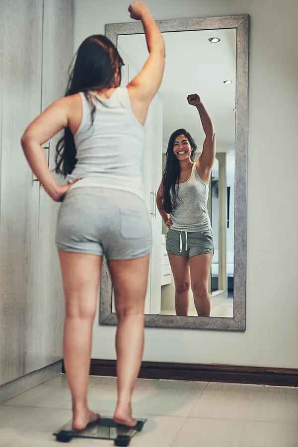 A woman in workout clothes stands on a scale looking in the mirror and pumping her hands in celebration of her successful Tirzepatide treatment from Brandi Terra, NP at Oak & Stone Wellness in San Diego.